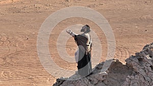 A man with a staff in his hand and the clothes of a shaman stands on a rock among the sands of the desert and dances