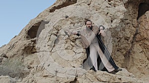 A man with a staff in his hand and the clothes of a shaman stands on a rock among the sands of the desert and dances