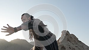 A man with a staff in his hand and the clothes of a shaman stands on a rock among the sands of the desert and dances