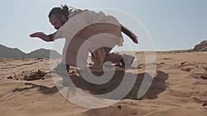 A man with a staff in his hand and the clothes of a shaman stands on a rock among the sands of the desert and dances