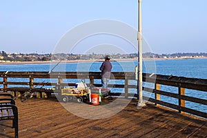 A man standing on the edge of a brown wooden pier leaning on the railing fishing with vast deep blue ocean water