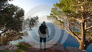 A man standing on the edge of a cliff and looking out to the sea at dawn.