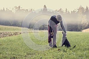 Man standing in green grassland bending down to pat his black do