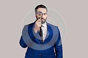 Man standing holding magnifying glass, looking at camera with big zoom eye, verifying authenticity.