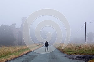 A man standing in the middle of the road next to a old ruined building on a moody misty morning