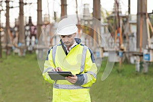 Man standing near high voltage substation and typing on device. Male engineer in uniform and helmet standing outdoor and