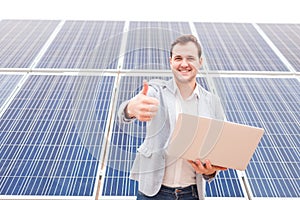 Man standing near solar panels, holding laptop, smiling and showing his index finger in front of him. Outdoors