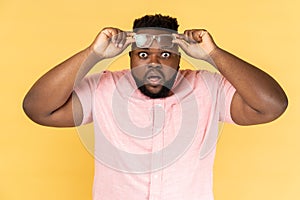 Man standing raised his optical glasses and looking at camera with big eyes and open mouth.