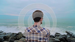 Man is standing on the rocky shore, looking at stormy sea waves