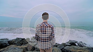 Man is standing on the rocky shore, looking at stormy sea waves: slow motion