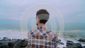 Man is standing on the rocky shore, looking at stormy sea waves: slow motion