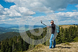 A man standing on the top of high hill with action camera - making selfie, high in mountains. beautiful nature and clouds with blu