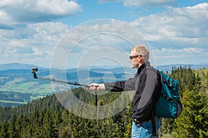 A man standing on the top of high hill with action camera - making selfie, high in mountains. beautiful nature and clouds with blu