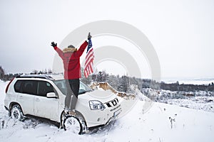 Man standing on the top of the hill with usa flag in his hands near suv car looking for beautiful winter view