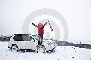 Man standing on the top of the hill with usa flag in his hands near suv car looking for beautiful winter view
