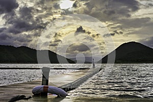 Man standing on a wooden pathway on the water, background with mountains and gloomy skies. The Srinakharin Dam