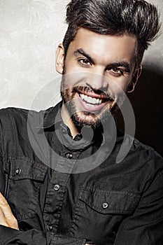 A man stands against the wall and crosses his arms over his chest in a black shirt his face expresses laughter and joy