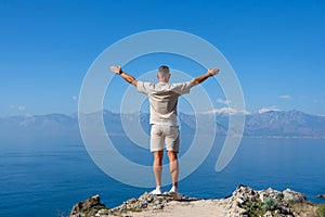 Man stands with arms raised on rock blue ocean mountain peaks horizon. Active holiday adventure, tourism action, healthy summer