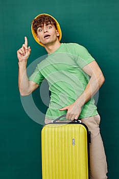 man stands on a background, dressed in a yellow hat, holding a bright travel suitcase in his hand, looking pleasantly