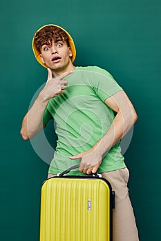 man stands on a background, dressed in a yellow hat, holding a bright travel suitcase in his hand, looking pleasantly