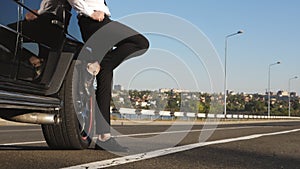 A man stands at a black car, leaning on the wheel with his foot. Waiting for a tow truck on the highway.