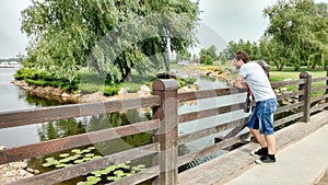 A man stands on the bridge and looks at the view: a pond, water lilies, an island with a willow, a neatly trimmed lawn