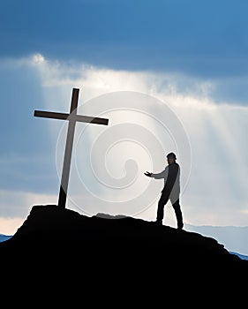 Man stands before a cross on a mountain, engaging in prayer with his hands folded