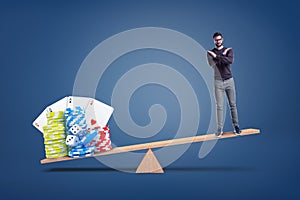 A man stands making a cross gesture with his arms while standing on a seesaw with heavier casino chips on another side.