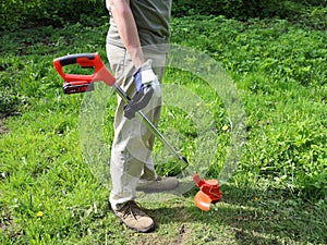 Man stands on meadow and holds an electric trimmer for mow grass, on sunny day