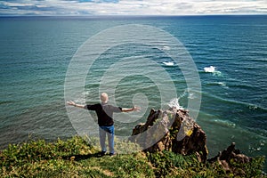 Man stands with open arms on cliff in front of Pacific ocean