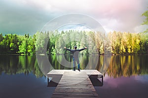 Man stands on the  pier of a beautiful lake,  Young man enjoying life, his arms open, a rainbow over the lake.
