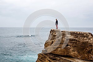 Man stands on a sea cliff, looking into the distance on a dark stormy sea.