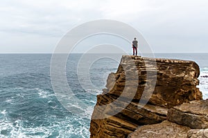 Man stands on a sea cliff, looking into the distance on a dark stormy sea.