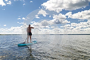 A man stands on a SUP board with a paddle in the lake on a sunny day against the background of white clouds.