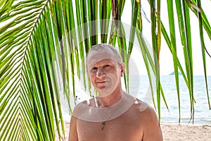 A man stands under a palm tree near the sea