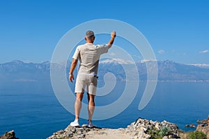 Man stands waving hand greeting on rock blue ocean mountain peaks horizon. Active holiday adventure, tourism action, healthy