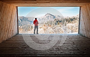 Man stay in big wooden hangar and looks on snowy mountains