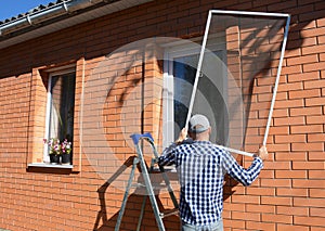 A man on a step ladder is installing a mosquito net, fly screen or frame on plastic window  to prevent different insects, bugs