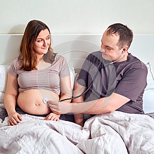 A man with a stethoscope and a pregnant wife in bed. Pregnant woman with her doctor husband in bed