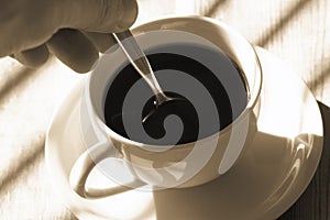 Man stiring a cup of black coffee in sunlight on a wooden table