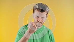 A man with stubble stands against a yellow wall and shakes his fist at the camera. A man in a green T-shirt shows his