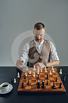 Man with stylish mustache, dressed in brown vest, white shirt is playing chess. Nearby is a cup of coffee. Grey