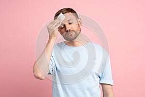 Man suffering from heat stuffiness wiping sweat on forehead with paper napkin isolated on pink wall