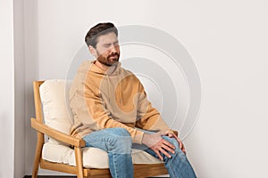 Man suffering from leg pain and touching knee on soft armchair indoors, space for text