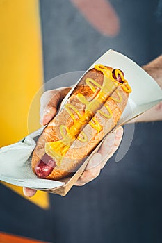 Man suggesting freshly prepared hotdog with cheese in a paper box. Food delivery or take awat food concept. Yellow background