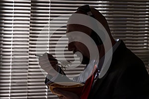 A man in a suit and headphones eats pasta against the background of a window with blinds. A special agent`s lunch at work, wiretap