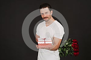 Man in suit is posing on black background with gift box and red roses in hands