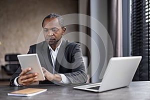 A man in a suit is sitting at a desk with a tablet and a laptop