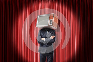 Man in suit standing with arms folded, with TV set instead head, in spotlight near red stage curtain.