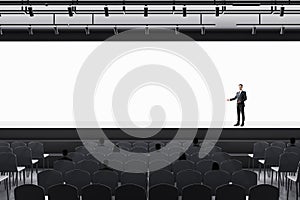 Man in suit standing in modern grey auditorium with seating and empty screen with mock up place for your advertisement. Show